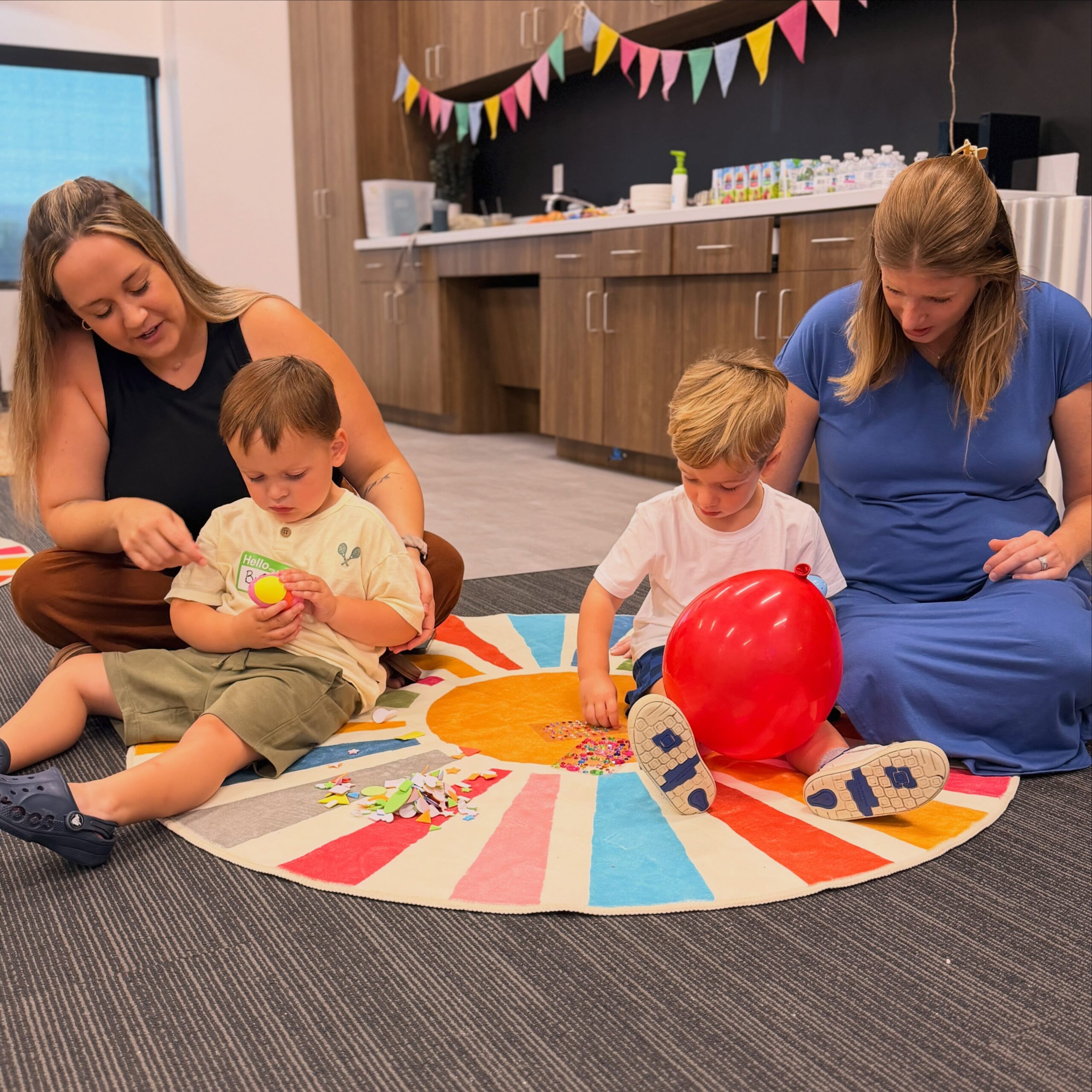Adults help young children with a craft activity on a colorful rug.