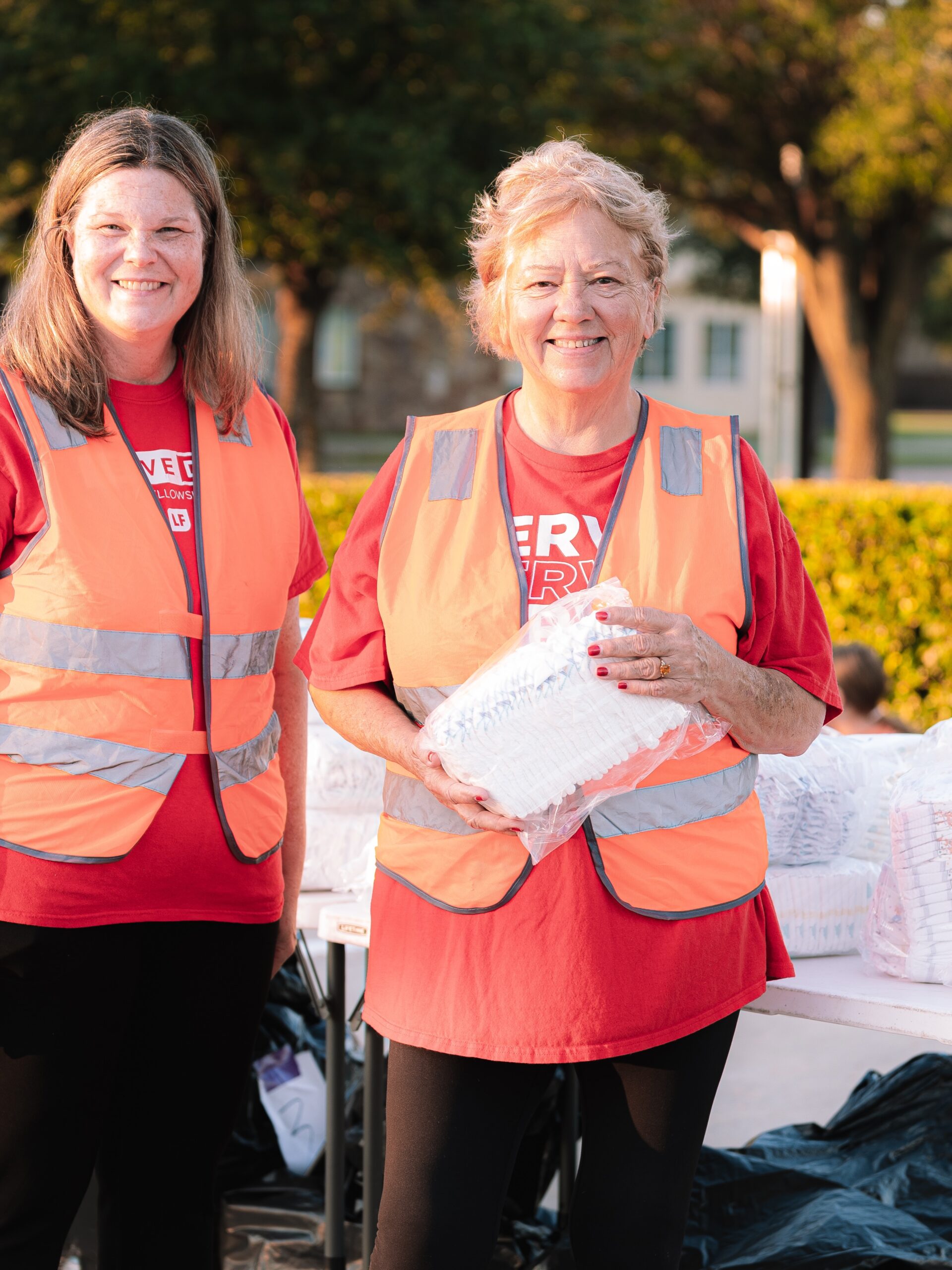 Two smiling volunteers in reflective vests stand together behind an outreach table.